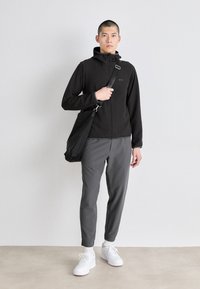 Črna jakna na zadrgo s kapuco, sivi slim-fit hlače in bele superge. Oseba drži črno torbo preko ramena.