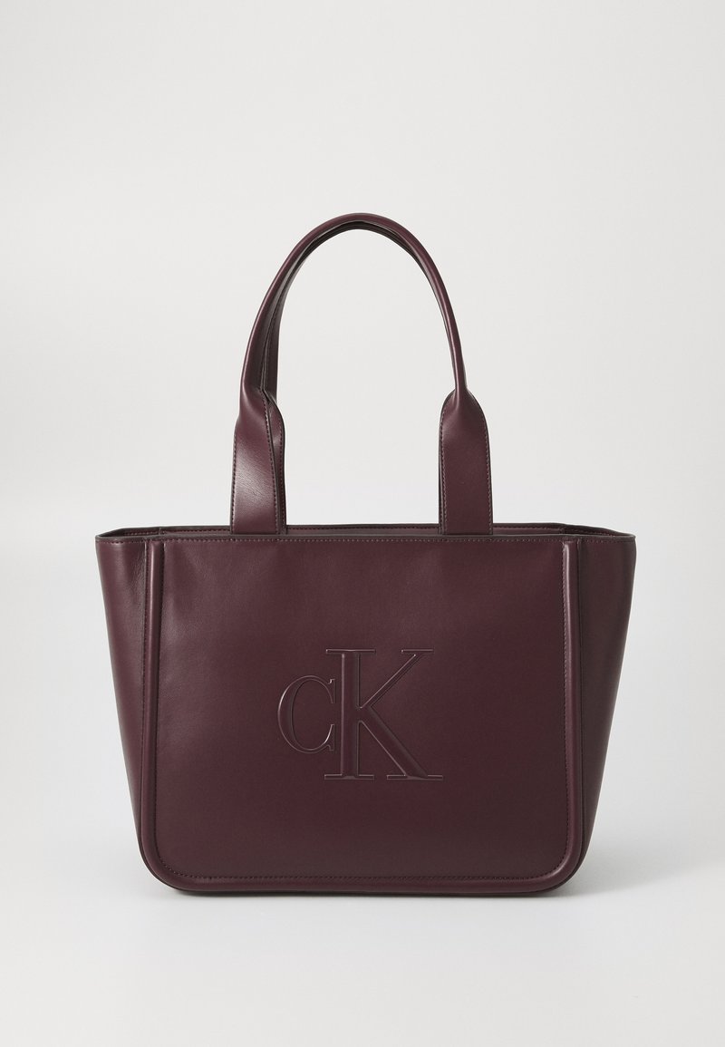 Burgundy leather tote bag with two top handles, featuring an embossed cK logo on the front and clean, structured design. Smooth texture.