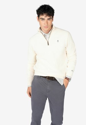 Suéter crema con cremallera y textura acanalada, combinado con una camisa de rayas y pantalones grises. Incluye un sutil logo en el pecho.