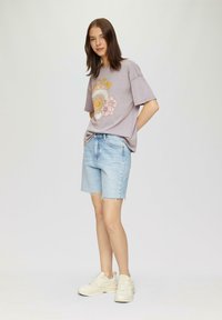 T-shirt graphique violet avec design floral, associé à un short en denim bleu clair avec un ourlet effiloché. Chaussures de sport claires.