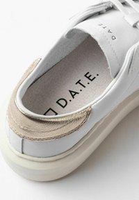 Zapatilla blanca de cuero con un acento metálico dorado en el talón, plantilla texturizada y logotipo "D.A.T.E." impreso en negro.