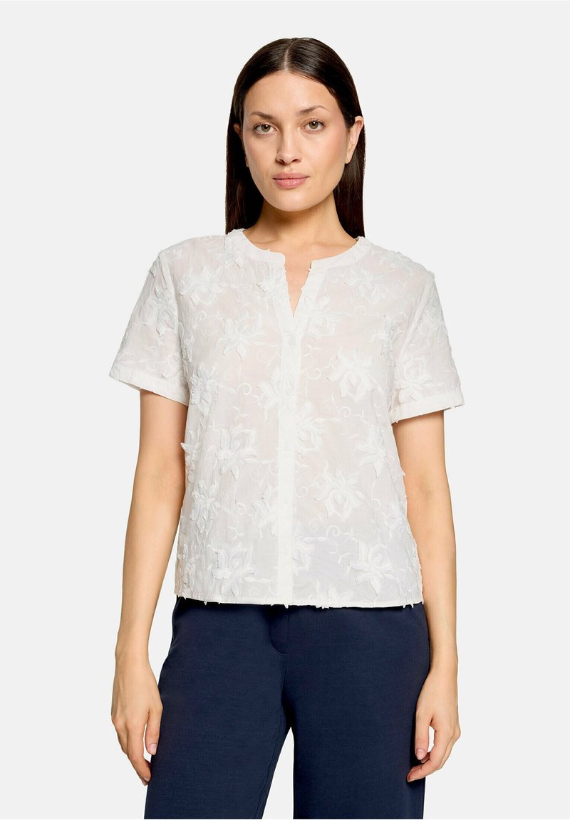 Femme aux cheveux foncés et raides porte un chemisier blanc à manches courtes orné de broderies florales et un pantalon bleu marine, debout devant un fond uni.