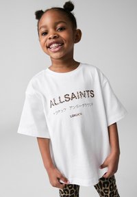 Biała, oversize'owa koszulka z napisem "ALLSAINTS" w leopardzie wzór, zestawiona z legginsami w czarno-brązowy leopardzi wzór. Gładki, bawełniany materiał.