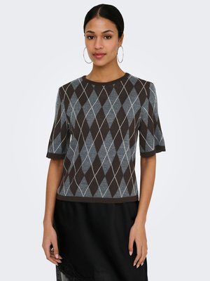 Junge Frau trägt einen kurzärmeligen, braun-grauem Rautenmuster-Strickpullover und einen schwarzen Rock, steht vor einem schlichten Hintergrund.