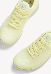 Paire de chaussures de sport jaune pâle avec dessus en mesh, lacets et semelles blanches, vues de dessus sur fond blanc.