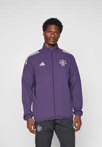 Lila Adidas-Jacke mit schwarzen Seitenakzenten, durchgehendem Reißverschluss, Stehkragen und Manchester United-Logo auf der Brust. Mit weißen Streifen an den Ärmeln.