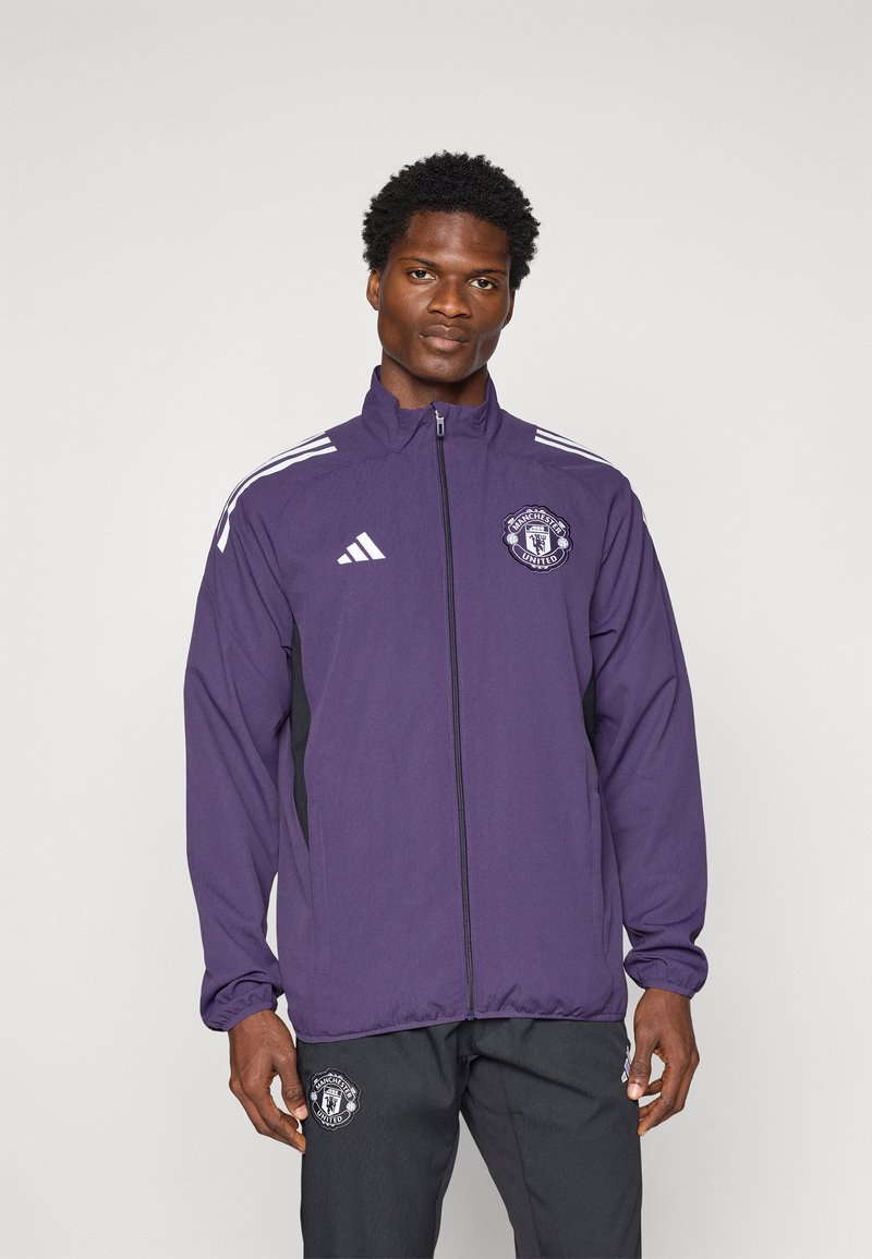Lila Adidas-Jacke mit schwarzen Seitenakzenten, durchgehendem Reißverschluss, Stehkragen und Manchester United-Logo auf der Brust. Mit weißen Streifen an den Ärmeln.