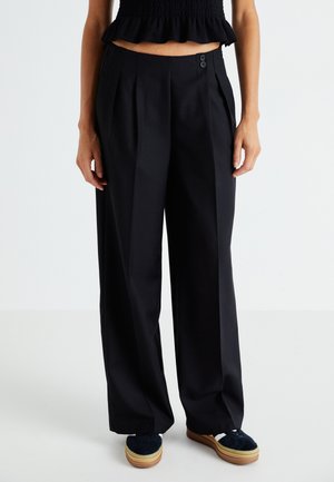 Svarte, vide bukser med legg, som har to knapper i midjen. Glatt stoff med skreddersydd design. Brukt med marineblå sneakers.