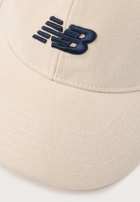 Beige čepice vyrobená z texturované látky, která na sobě má vyšívané logo "NB" v námořnické modré barvě a kontrastní bílé švy podél kšiltu.