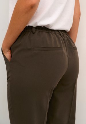 Personne portant un pantalon marron foncé à taille élastique avec des poches passepoilées à l'arrière et une chemise blanche rentrée, main reposant dans la poche latérale.