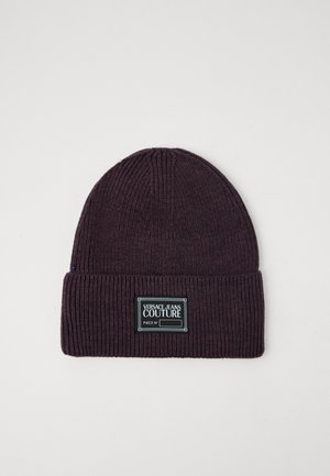 Sötétlila bordázott kötött sapka, hajtott szegéllyel. További jellemzői között szerepel egy téglalap alakú címke "VERSACE JEANS COUTURE" felirattal és díszvarrással.