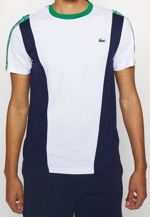 Mężczyzna w białej koszuli z krótkim rękawem z granatowymi panelami, zielonym kołnierzem i logo na piersi, stojący z ramionami swobodnie opuszczonymi wzdłuż ciała.