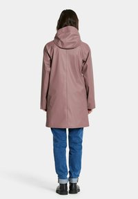 Ljusrosa vattentät kappa med huva, med rak skärning och långa ärmar, kombinerad med blå jeans och svarta skor.