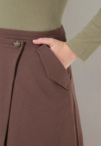 Bruine katoenen rok met een hand in een zak, voorzien van een decoratieve knoop en een gestructureerde top. De top is olijfgroen en geribbeld.