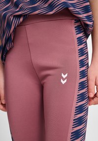 Różowe legginsy o gładkiej fakturze, z bocznymi panelami w falisty wzór w ciemnym niebieskim oraz białym logo w kształcie chevronu na udzie.