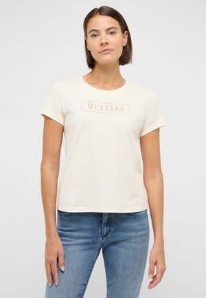 Junge Frau in einem cremefarbenen T-Shirt mit Mustang-Logo und blauer Jeans steht vor einem schlichten hellen Hintergrund.