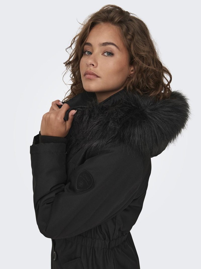 Parka noire avec une capuche doublée de fourrure, taille cintrée et poignets élastiques. Présente un insigne sur le bras et un tissu lisse et texturé.