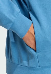 Felpa blu con maniche lunghe, polsini a coste e una tasca laterale. Il tessuto ha una texture morbida, che presenta un design liscio e casual.