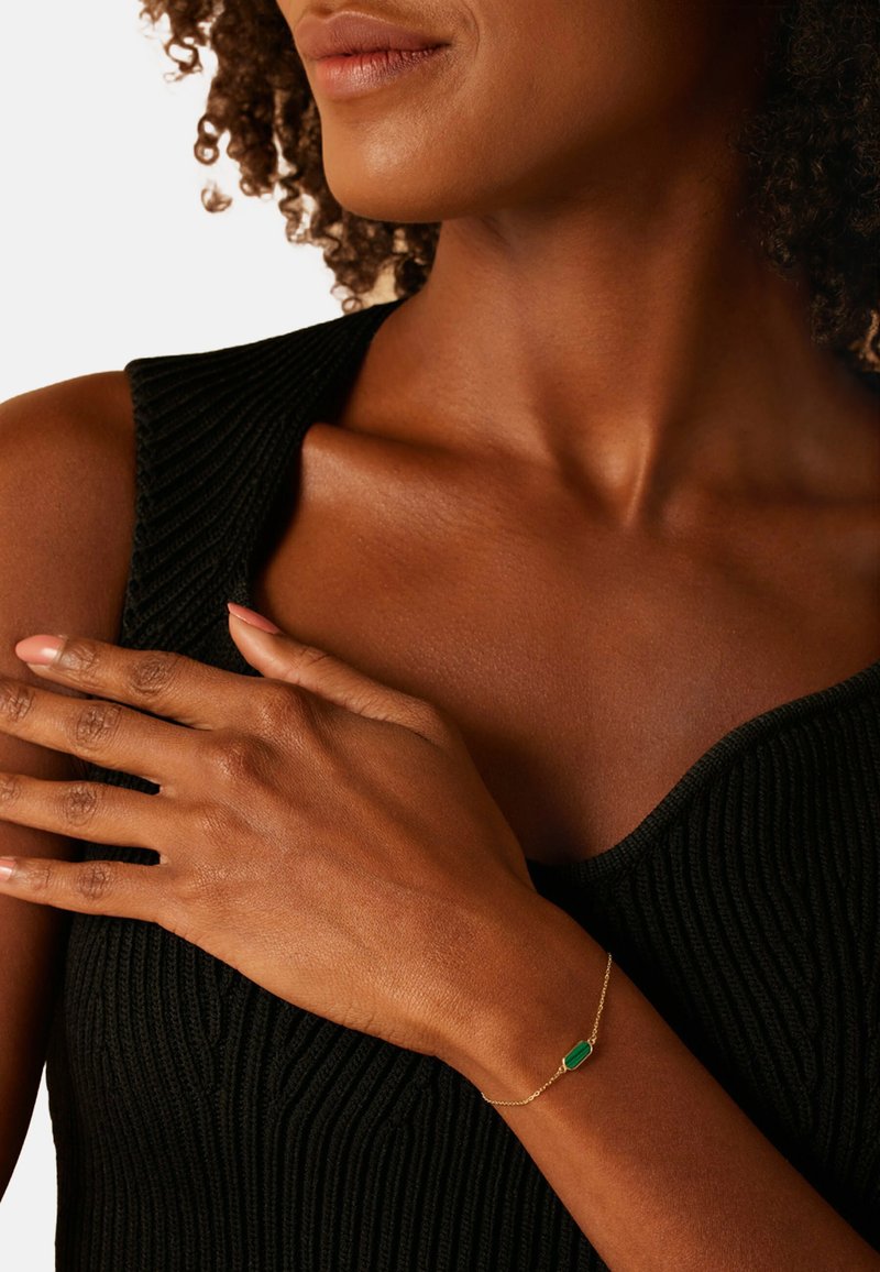 Gouden armband met een smaragdgroene rechthoekige steen bevestigd aan een fijne ketting, gedragen aan een hand tegen een gestructureerd zwart top.