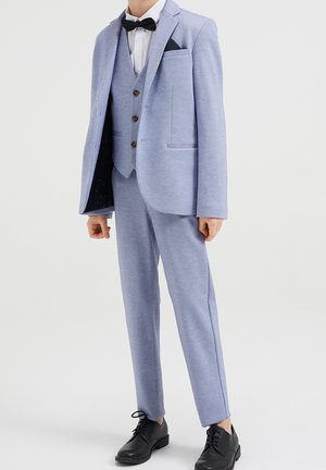 Niño vestido con un traje de tres piezas azul claro, con pajarita negra y zapatos de vestir negros, de pie contra un fondo liso.