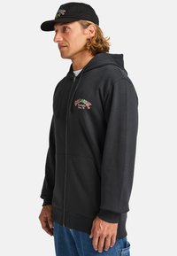 Zwarte ritssluiting hoodie met een kangoeroezak, met meerkleurige borduursels van "Billabong Australia" op de borst en geribde mouwen.