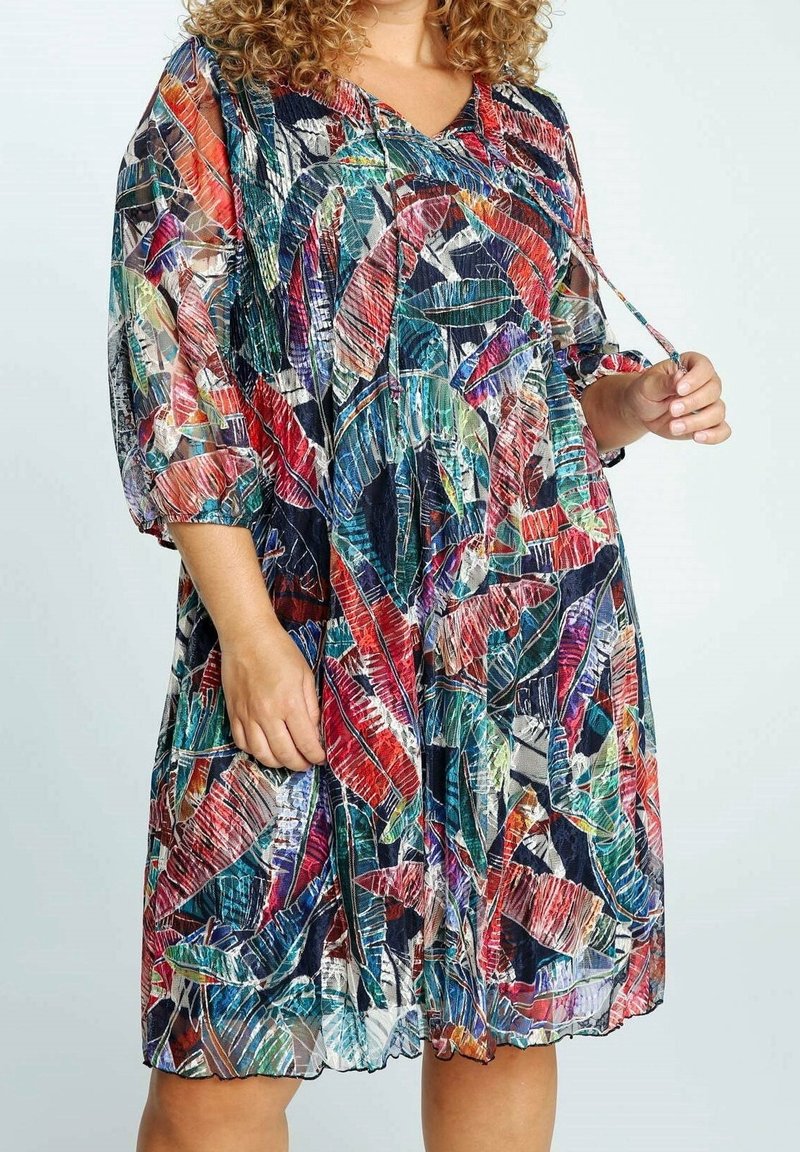 Nainen, joka pukeutuu polvipituiseen mekkoon, jossa on värikäs abstrakti lehtikuvio, kolmivartiset hihat ja nyöritys kaula-aukossa.