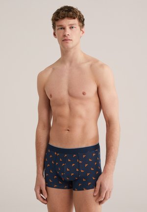 Muž stojaci na neutrálno sfarbenom pozadí, oblečený v námorníckych boxerkách s vzorom oranžových croissantov, pozerá priamo pred seba.