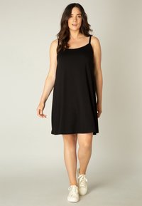 Zwarte mouwloze jurk met een ronde halslijn, vloeiende stof en een losse pasvorm. Vergezeld van witte sneakers voor een casual uitstraling.