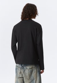 Cămașă neagră cu mânecă lungă din bumbac, cu guler rotund. Prezintă o croială relaxată și detalii subtile de cusătură. Purcată cu blugi din denim albastru deschis.