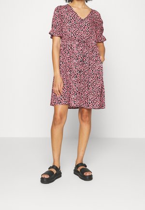 Mujer con un vestido corto de flores rosa y negro con mangas abullonadas y sandalias negras de plataforma, posando frente a un fondo blanco.