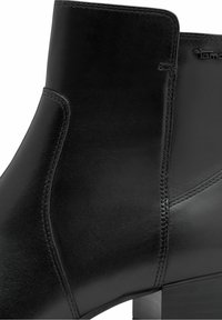 Gros plan sur une botte noire en cuir à la cheville, présentant une texture lisse, des coutures visibles et un talon carré bas.