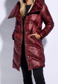 Manteau rembourré bourgogne avec un col haut, fermeture éclair et poches. Présente une texture brillante, matelassée et un motif de couture en chevrons.