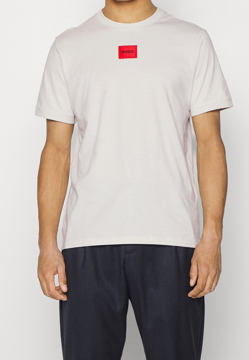T-shirt gris clair en coton, avec un logo carré rouge portant l'inscription "HUGO" au centre. Manches courtes et coupe décontractée. Porté avec un pantalon foncé.