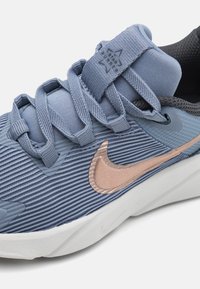 Világoskék sportcipő bordás textúrával, szintetikus felsőrésszel és rozé arany Nike logóval. Szürke részletekkel és párnázott nyelvvel rendelkezik.