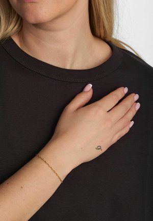 Czarna koszulka z okrągłym dekoltem, noszona przez osobę; ręka spoczywa na piersi, ozdobiona delikatną złotą bransoletką i małym tatuażem.