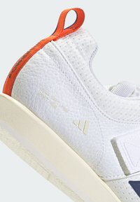White athletic shoes with textured material, orange accents, and a cream-colored sole. Features branding and a rear pull tab.