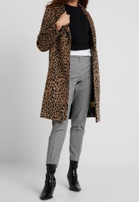 Abrigo tipo trench de estampado de leopardo en marrón y negro, hecho de material suave, combinado con pantalones de cuadros en gris y botines negros brillantes.