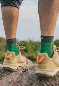 Zapatillas deportivas marrones con acentos naranjas, combinadas con calcetines verdes con estampado justo por encima del tobillo, llevadas mientras se está de pie sobre una roca.