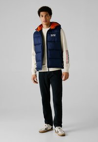 Navy plava prsluk s narančastim ovratnikom, visokim ovratnikom, prednjim patentnim zatvaračem i kontrastnim logotipom. Nošen preko svijetlog džempera i tamnih traperica.