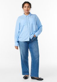 Sieviete ar ķermeni matiem stāv, tērpta gaismas zilā krekliņā ar pogām, zilos džinsos un melnos kājāmgājēju apavos uz baltas fona.