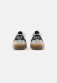 Nike sko med en hvid og lys grå overdel, sorte accenter og en brun gummisål, hvor "NIKE" logoet er tydeligt vist på hælen.