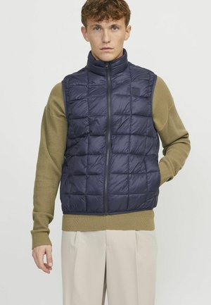 Veste isolée marine avec une fermeture éclair à l'avant, présentant un design matelassé. Associée à un pull texturé vert olive et un pantalon beige clair.