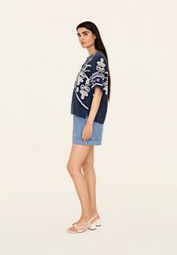 Mörkblå topp med vita broderade blommönster, korta ärmar, i kombination med ljus denimshorts och ljusrosa klacksandaler.
