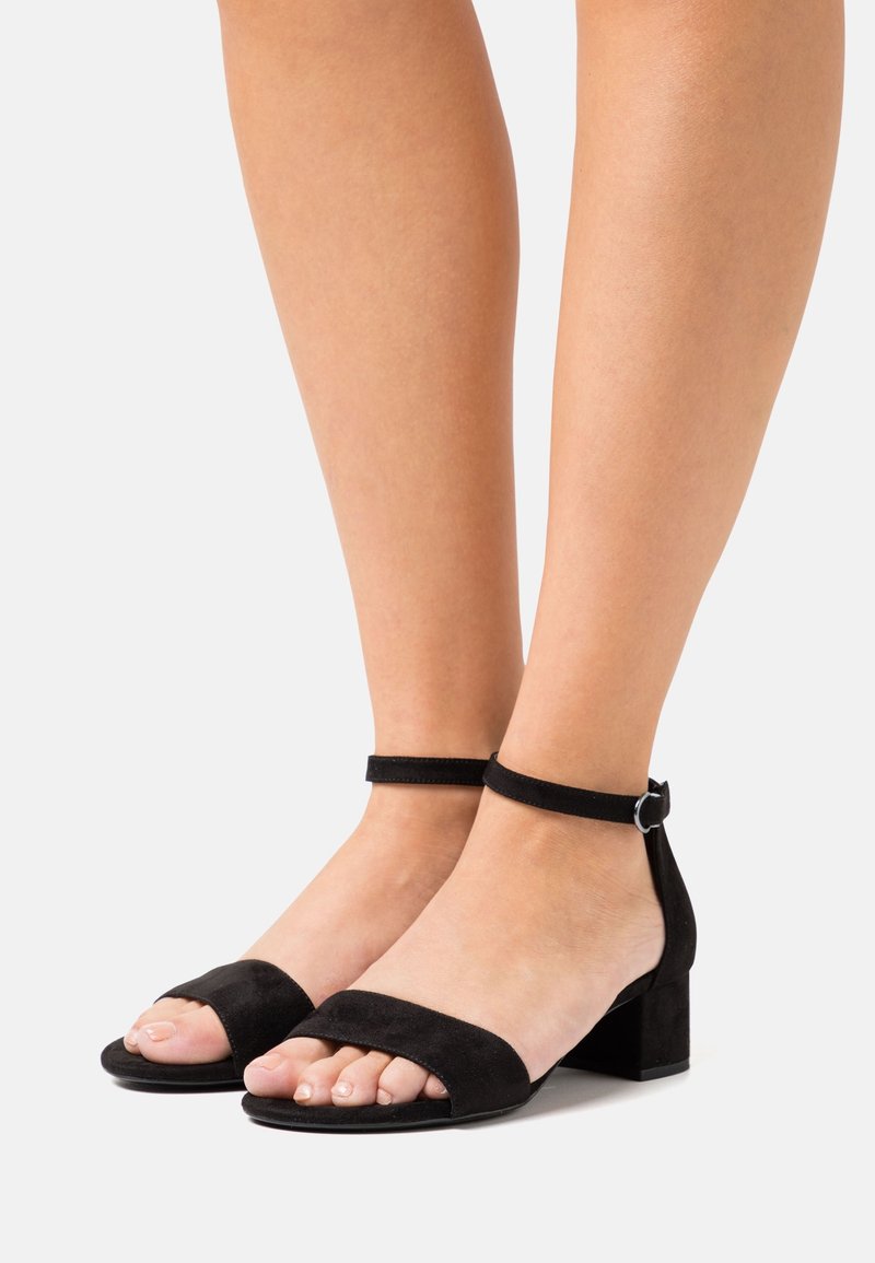 Pieds portant des sandales à talon bloc en daim noir, à bout ouvert, avec bride à la cheville et boucle sur fond blanc.