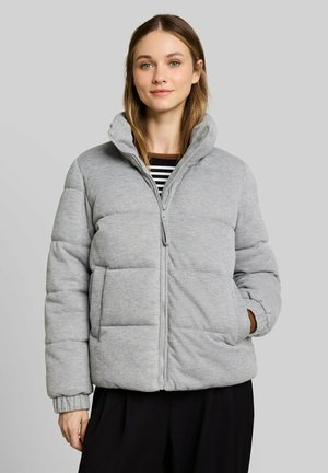 Junge Frau mit hellbraunen Haaren, die eine hellgraue gesteppt Jacke und schwarze Hose trägt, steht vor einem einfarbigen grauen Hintergrund.