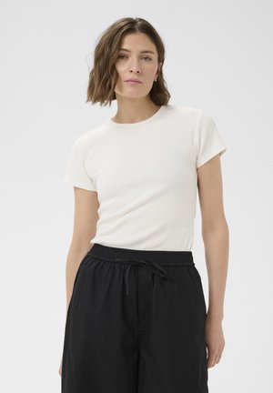 Vrouw met kort golvend bruin haar, gekleed in een wit kortärmelig shirt en zwarte broek met aantrekkoord, tegen een effen lichte achtergrond.