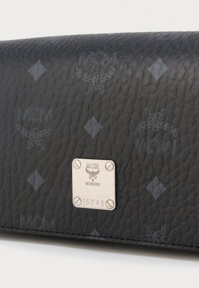 Μαύρη υφασμάτινη δερμάτινη επιφάνεια με επαναλαμβανόμενα λογότυπα MCM και ασημένια μεταλλική πλακέτα χαραγμένη με το MCM München και τον αριθμό 15248.