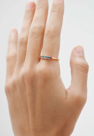 Mano con un anillo de oro que presenta cinco pequeñas gemas en azul y rojo, llevado en el dedo medio sobre un fondo claro y liso.