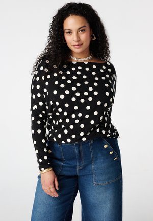 Vrouw met krullend haar, gekleed in een zwarte top met lange mouwen en witte stippen, en een blauwe hooggetailleerde spijkerbroek met gouden knopen, poserend met zelfvertrouwen.