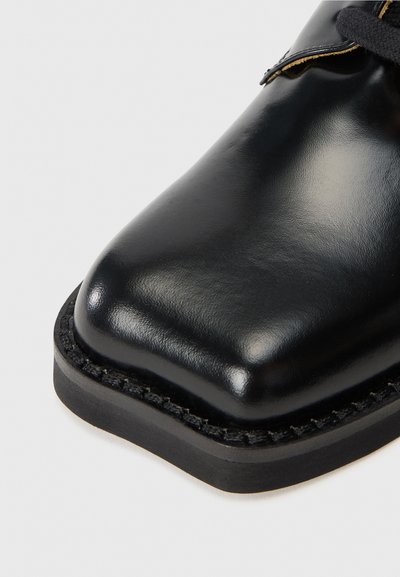 Svart lädersko med en slät, blank finish. Rundad tåform och synlig söm längs kanten av gummisulans.